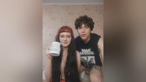 Media: Video of a pale-skinned, long-haired woman with red hair and glasses, holding a white mug, and a curly-haired, shirtless man with tattoos, wearing a black \"Slayer\" t-shirt, seated indoors with beige wallpaper.