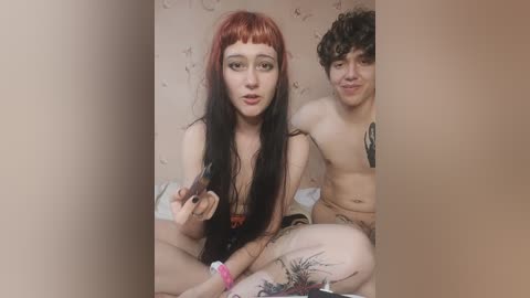 Media: Video of two young people with pale skin and tattoos. The girl with long red hair and the boy with curly hair sit close on a bed, both topless, holding a smartphone.