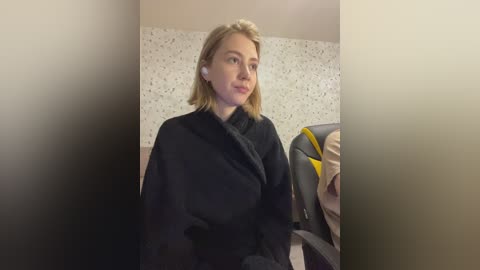 Video of a fair-skinned woman with blonde hair, wearing a black robe, sitting in a bathroom with speckled white walls. She holds a yellow and black hairdryer.