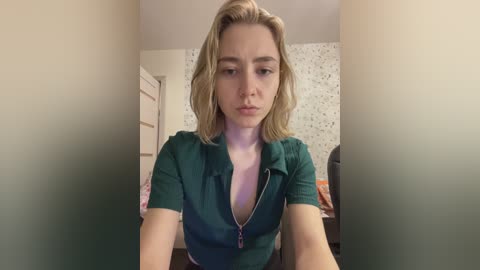 Video of a blonde woman with light skin, wearing a teal shirt, seated in a dimly lit room with floral wallpaper.