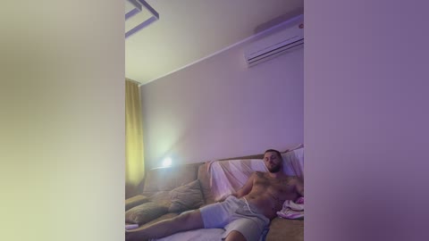 Video of a shirtless, muscular man with short brown hair, wearing light gray shorts, lying on a beige couch in a dimly lit room with a yellow curtain, an air conditioner, and a lamp.