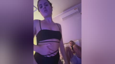 A video shows a young woman in black lingerie, with a man in a white tank top in the background, in a dimly lit room with purple lighting.
