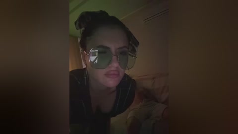 A dimly-lit video of a woman with light skin, wearing oversized sunglasses and a black top, bending forward, taken from a low angle. The background shows a beige wall and a bed with patterned sheets.