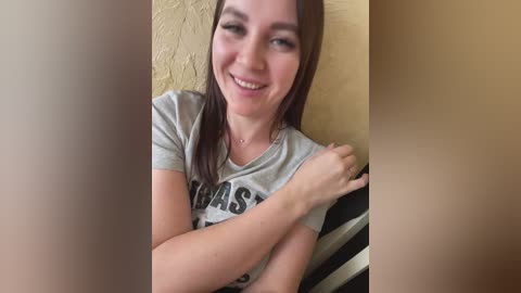 Media: Video of a smiling young woman with fair skin and brown hair, wearing a grey t-shirt with \"CORNELIUS\" in black letters, sitting on a striped pillow against a textured yellow wall.