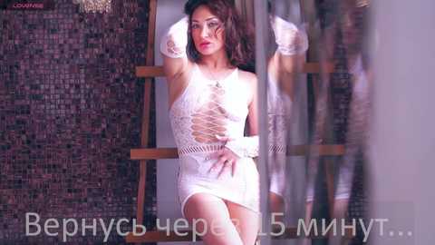 Media: Video of a curvaceous woman with fair skin, long dark hair, and full lips, wearing a revealing white lace mini dress, posing seductively in a dimly lit room with mosaic tiled walls. Russian text at the bottom.