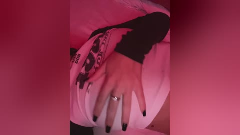 Media: Video of a person's hand with black nail polish, wearing a white shirt with black and white graphic print, partially covered by another person's arm, in a dimly lit, pink-lit room.