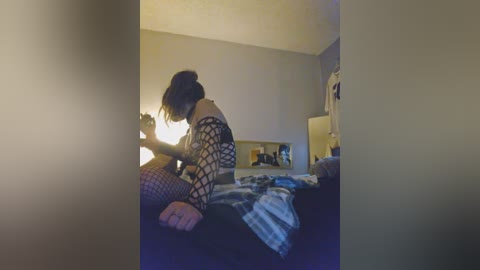 A video captures a young person, likely a teenager, sitting on a bed with a messy room in the background. The person is wearing a black, fishnet top and has their hair tied back. The lighting is dim, creating a moody atmosphere.