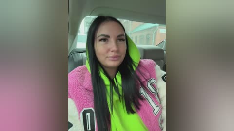 Video of a young woman with long, straight black hair, wearing a neon green and pink furry jacket with a large white \"1\" on the sleeve, seated in a car.