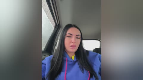 A video of a young woman with long black hair, wearing a blue jacket and a yellow shirt, sitting in a car with a gray interior.