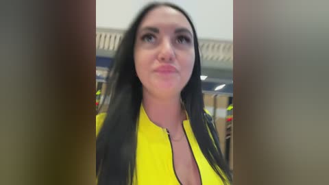 A video of a woman with long black hair, fair skin, and a slim physique, wearing a yellow jacket. The background shows an indoor setting with blurred details of people and a ceiling.