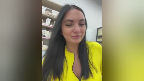 A video of a smiling woman with long, straight black hair, wearing a bright yellow blouse, standing in a pharmacy with shelves of medical products in the background.