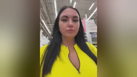 Video of a woman with long black hair, wearing a bright yellow zip-up hoodie, standing in a dimly lit, industrial warehouse setting with exposed pipes and fluorescent lights.
