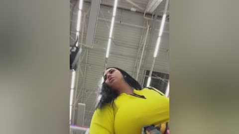 Video of a curvaceous woman with long black hair wearing a bright yellow top, captured from a low angle in a warehouse with fluorescent lighting and metal shelving.