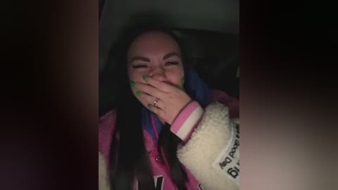 Video of a young woman with light skin, long black hair, and bright green nails, wearing a pink and white jacket, covering her mouth, against a dark background.