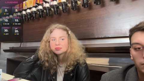 Media: Video of a blonde woman with curly hair, wearing a black leather jacket, seated at a bar counter. Background shows a liquor display and a man partially visible on the right.