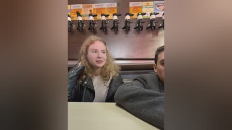 Media: Video of a young woman with long, wavy blonde hair, wearing a black leather jacket and white top, sitting at a booth in a diner. A sign with a cartoon chef hangs above, and another person is partially visible beside her.
