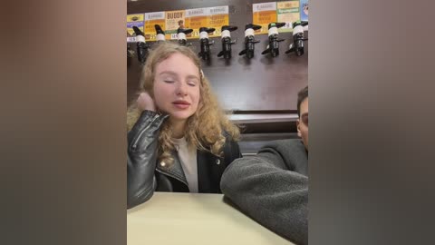 Media: Video of a young blonde woman with curly hair, wearing a black leather jacket, resting her head on her hand, seated in a dimly lit caf\u00e9 with a busy counter in the background.