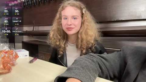 Media: A video of a young, fair-skinned woman with curly blonde hair and a light jacket, sitting at a counter with a sandwich and a box of tissues, set in a dimly lit room.