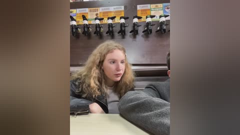 Media: A video of a young woman with long, wavy blonde hair, wearing a black jacket and grey pants, seated in a fast-food restaurant. In the background, a row of black coffee machines and a wall of promotional posters is visible.