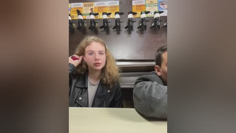 Media: Video of a young blonde girl with curly hair, wearing a black leather jacket, sitting at a table in a diner with a motorcycle display in the background.