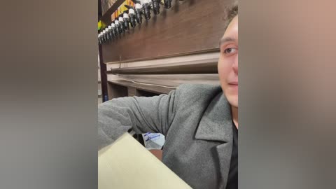 Media: A video of a young man with fair skin, short brown hair, and a gray blazer, partially obscured by a blurred background featuring a wooden cabinet and tools.