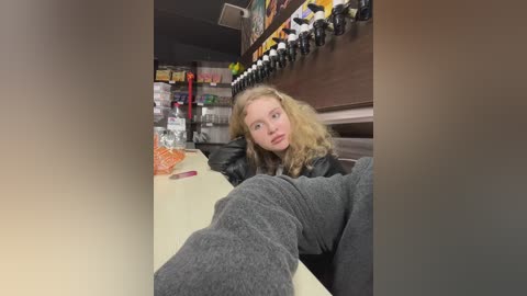 Media: Video of a blonde girl in a gray sweater, leaning on a table in a cluttered, dimly-lit room with shelves of toys and a black pinball machine in the background.