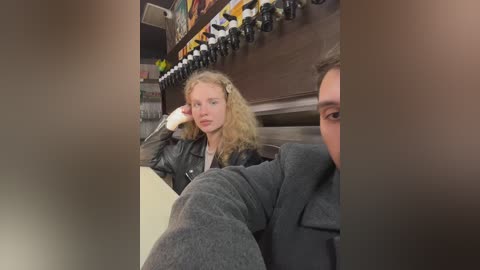 Media: Video of two people in a bowling alley. A woman with curly blonde hair, wearing a black jacket, sits on a bowling lane, resting her head on her hand. A man with short brown hair, dressed in a gray sweater, stands behind her, partially out of focus.