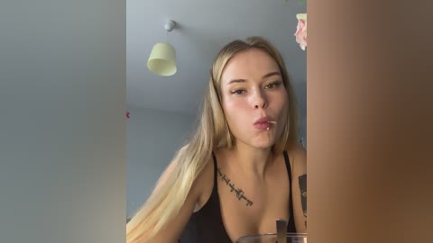 Media: Video of a young woman with long blonde hair, wearing a black top, blowing a kiss, against a minimalist gray wall with a pendant light and pink flower in the background.