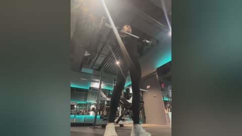 Media: Video of a young man in a gym, wearing a black hoodie, black pants, and white sneakers, lifting weights with a barbell. Background features modern gym equipment, teal lighting, and industrial-style decor.