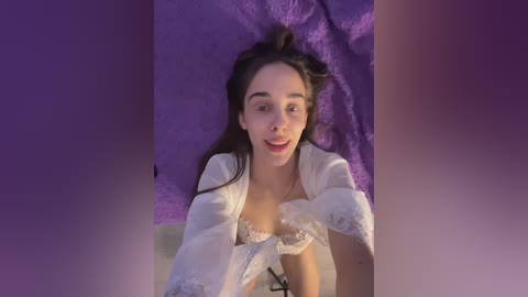 Media: Video of a young woman with long dark hair, fair skin, and a slender build, lying on a purple blanket. She wears a white lace bra and robe, smiling at the camera.