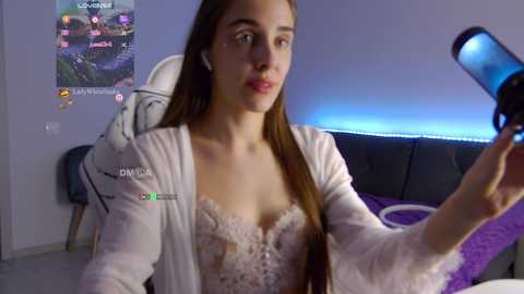 Media: Video of a young woman with long brown hair, wearing a white robe and lace bra, recording herself on a gaming chair in a dimly lit room with a gaming setup in the background.