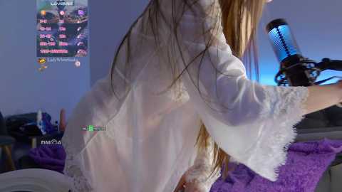 Media: Video of a slender, light-skinned woman with long blonde hair, wearing a sheer white lace robe, standing in a dimly lit bedroom with a purple blanket and blue lighting, streaming on Twitch.