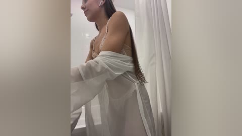 Media: A video of a slender, light-skinned woman with long, straight brown hair, wearing sheer white lingerie and an open white robe, standing in a dimly lit bathroom with white walls and a shower curtain.