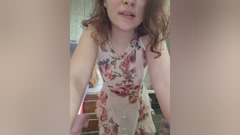 Media: Video of a Caucasian woman with curly brown hair, wearing a floral-patterned, sleeveless dress, standing in a kitchen with tiled walls and a sink visible.