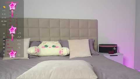 Media: Video of a modern bedroom with a beige tufted headboard, a plush white pillow, and a decorative cat pillow on a gray bed. A calendar with pink stars overlays the image, indicating a playful, whimsical setting.