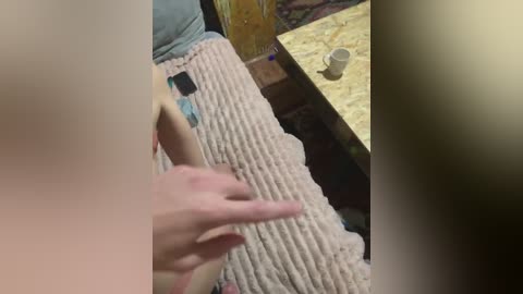 Video of a person with light skin, wearing a textured beige blanket, holding a black smartphone, on a wooden table with a beige cup, in a rustic room with exposed wooden walls.