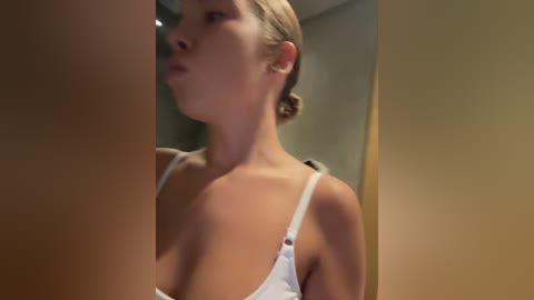 Media: A video of a light-skinned woman with blonde hair in a bun, wearing a white spaghetti strap top, blowing into a large brown bottle, taken indoors with blurred background.