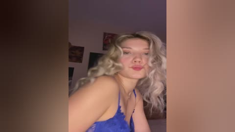 Media: Video of a blonde woman with wavy hair, wearing a blue lace lingerie top, posing provocatively in a dimly lit room with abstract wall art in the background.