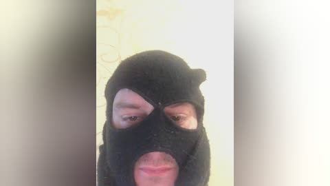 Video of a person wearing a black mask with two small eye holes and a large mouth hole, set against a light, textured background.