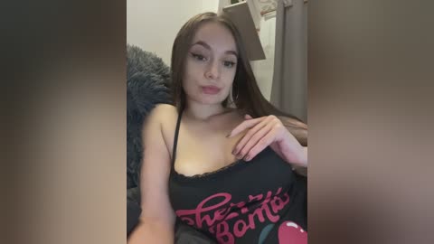A video of a young woman with long brown hair, wearing a black tank top with red text, sitting on a gray couch, indoors.