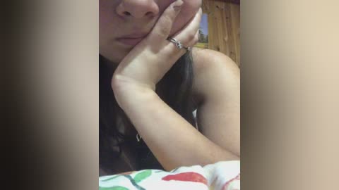 A close-up video of a woman's face with fair skin, resting her chin on her hand, wearing a black top, silver ring, and a colorful blanket.