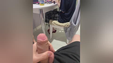 Media: Video of a person sitting at a table with a large erect penis in their hand, wearing a striped shirt and plaid shorts.