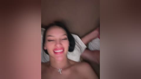 Media: A blurry, close-up video of a woman with long dark hair and light skin, smiling broadly, lying on her back with a white pillow behind her.