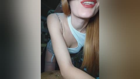Video of a smiling young woman with straight, long red hair, fair skin, and red lipstick, wearing a white tank top and patterned shorts, leaning over a table.