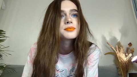 Video of a young, fair-skinned woman with long brown hair, wearing bright orange lipstick and a white tie-dye shirt. Background features a light wall, potted plants, and dried flowers.