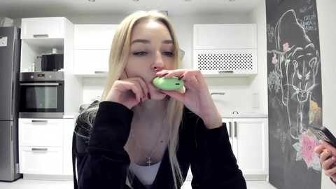 Media: Video of a blonde woman with fair skin, wearing a black jacket, eating a green apple in a modern white kitchen with black chalkboard wall.