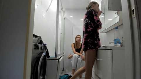 Media: Video of a young woman in a small, modern, white-walled laundry room with a woman sitting on a dryer. She wears a black tank top and white pants, and has light skin and long blonde hair.