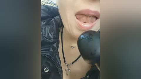 Video of a woman with pale skin, wearing a black leather jacket and a cross necklace, sticking out her tongue and speaking into a black microphone.