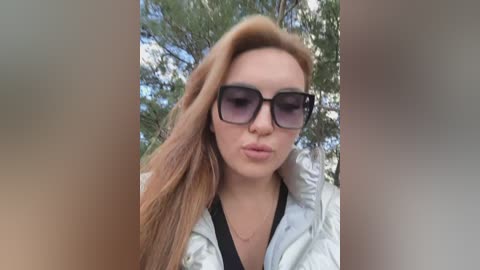 Video of a young woman with long, wavy, light brown hair, wearing large, black-framed sunglasses and a white puffer jacket, making a kissy face outdoors amidst green pine trees and blue sky.