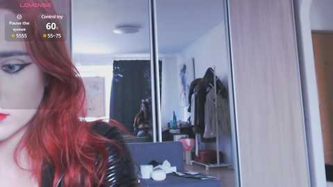 Video of a woman with long, wavy red hair, wearing black leather gloves, standing in front of a wardrobe mirror, reflecting a cluttered bedroom.